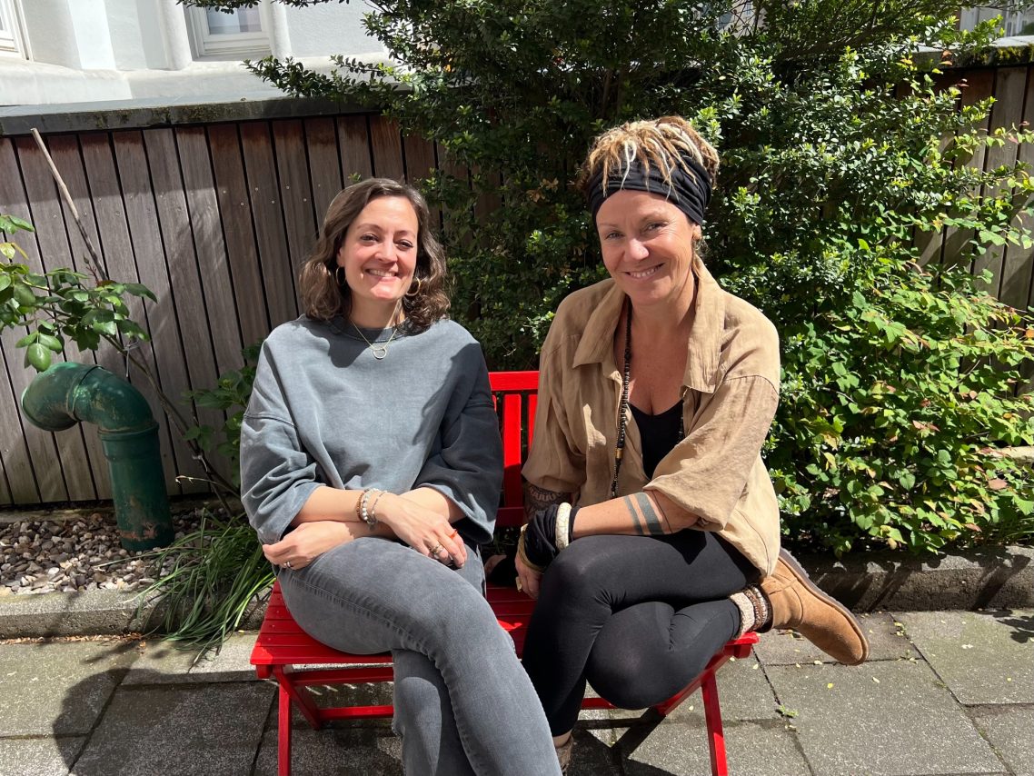 Zwei Frauen sitzen lächelnd auf einem roten Stuhl im Freien. Grüne Pflanzen im Hintergrund.